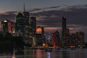 Brisbane city skyline at night, with tall buildings illuminated by lights, reflecting on the water below, under a moody sky.