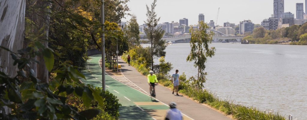 Scenic riverside bike and pedestrian path with trees, people walking, and city skyline in the background.