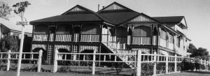Black and white photo of a traditional Queenslander-style house with a fenced yard.