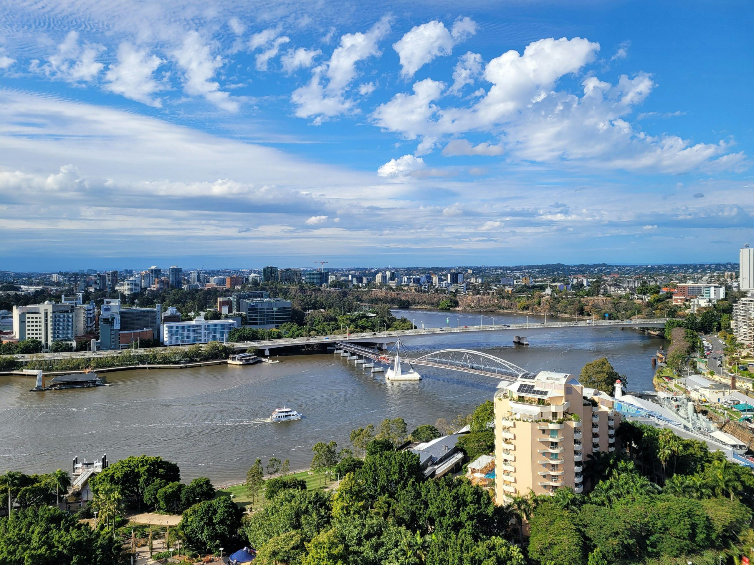 Aerial view of Brisbane River, city buildings, bridge, and greenery near Norman Park under a bright blue sky.