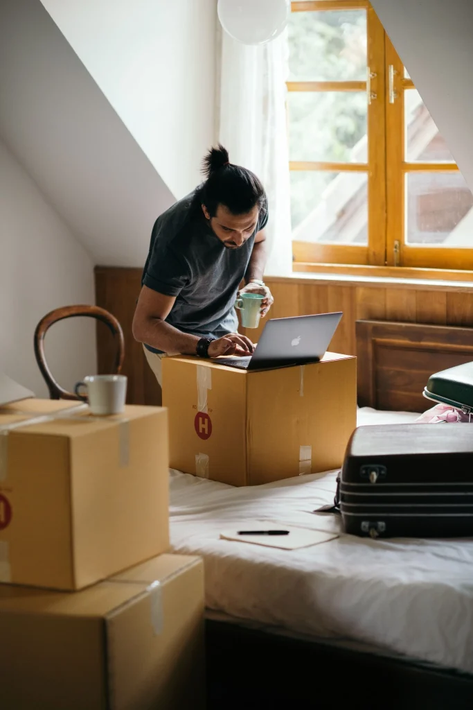 Man using a laptop among boxes in an Airbnb bedroom.