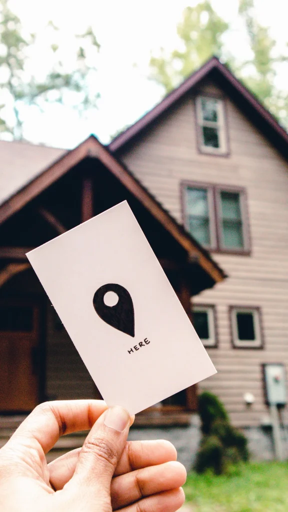 Hand holding a location card marked “Here” outside a house.